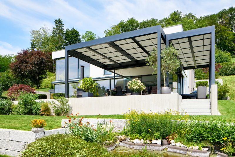 Referencia cubierta de patio independiente markilux markant con lona del toldo de luz adyacente a un edificio de cubo.