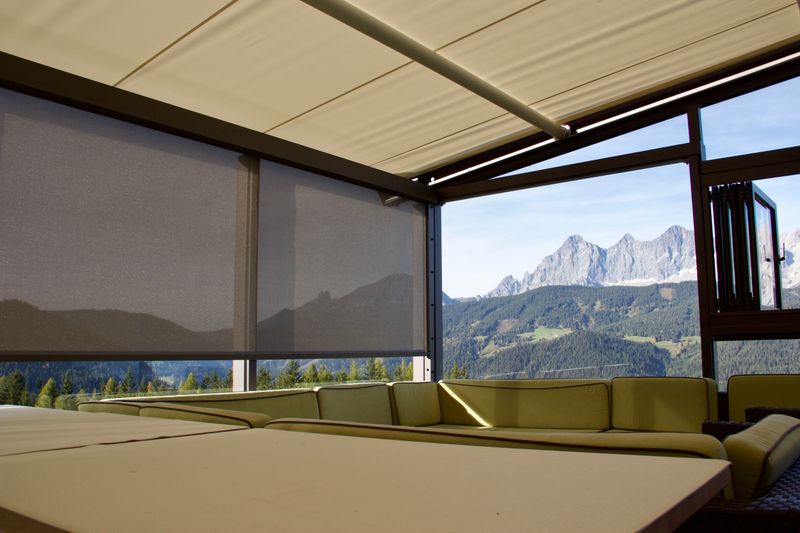 Vista desde el jardín de invierno de un prado alpino hacia los Alpes. Las ventanas están parcialmente sombreadas con el toldo vertical markilux 625, la vista se conserva, las montañas brillan a través del tejido de la lona.